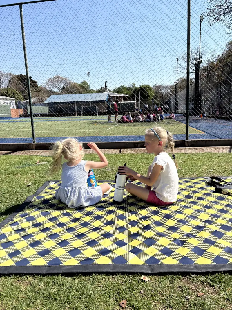 Two girls sitting on a yellow and navy checked picnic blanket on the grass beside a sports court